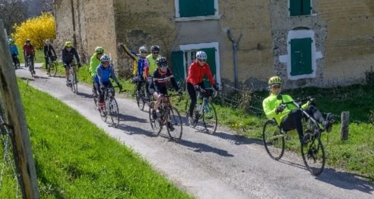 Une file de cyclistes du club sur une petite route de campagne, devant une ferme aux volets verts, au printemps.