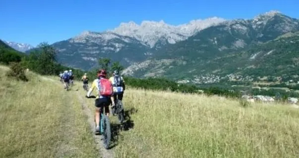 Vététistes du club en file sur un sentier herbeux, face à des crêtes calcaires enneigées.