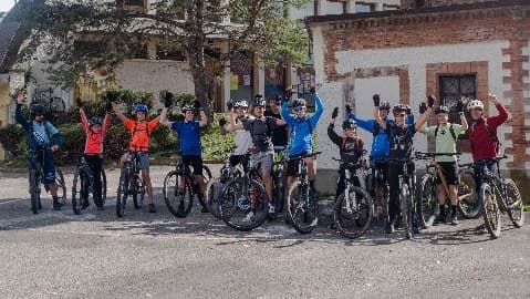 Une quinzaine de jeunes de la Team J, VTT à la main, bras levés devant un bâtiment en briques.
