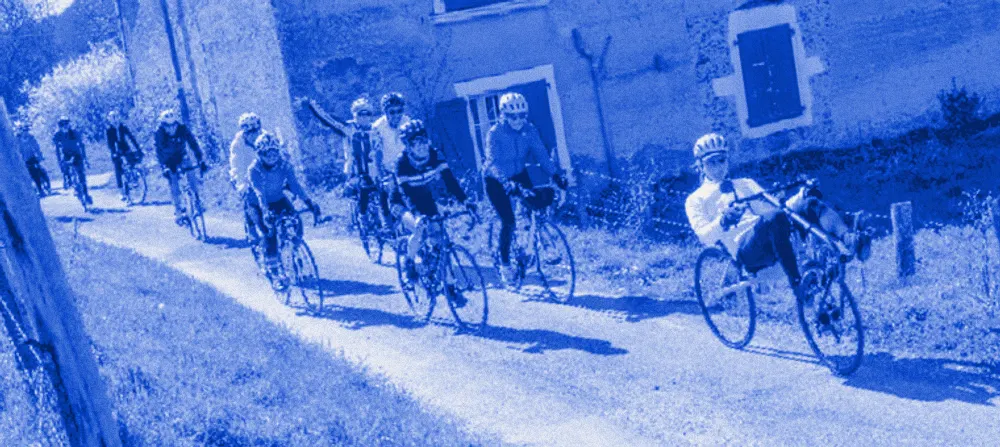 Un groupe de cyclistes sur une petite route le long d'une ferme en pierre, au printemps ; l'un d'eux roule en vélo couché.