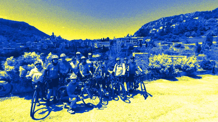 Un groupe du club pose avec ses vélos de route devant un village de pierre à flanc de colline, entre des genêts en fleurs.